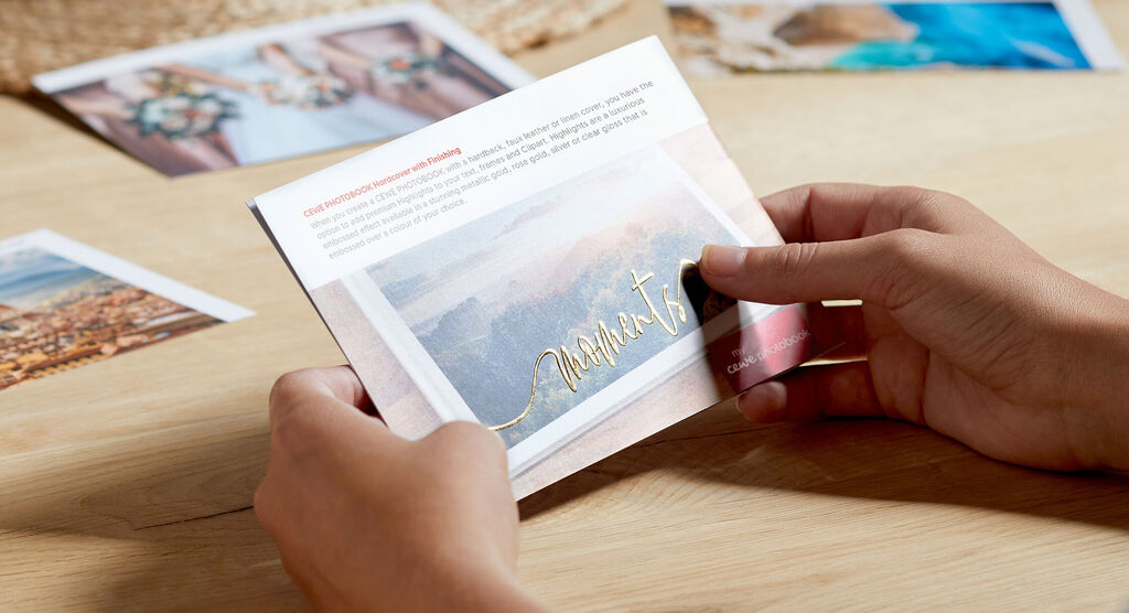 A person holds a CEWE photo book with a stunning landscape image and golden text, surrounded by more photos on a table.