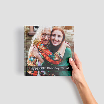 A person holds a CEWE photo book with 'Happy 60th Birthday Nana' written on it and is smiling.