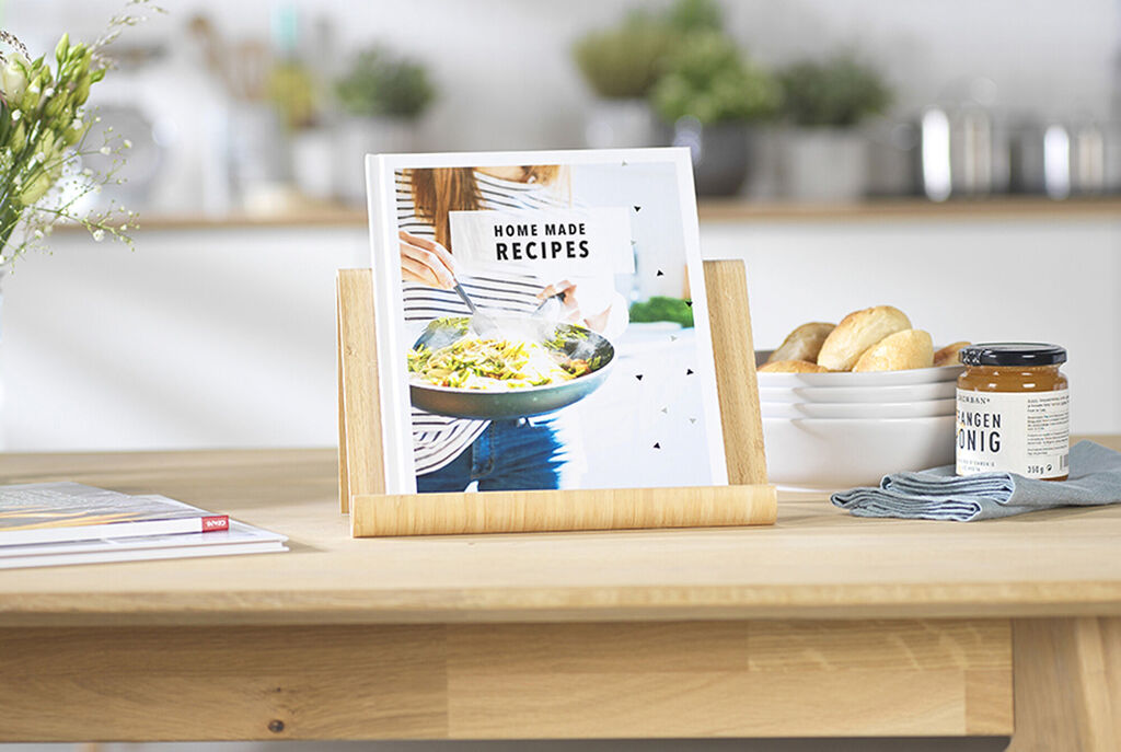 A lovingly designed recipe book is on a wooden stand. In the background, a bright kitchen is seen with a bowl, rolls, and a jar of jam.