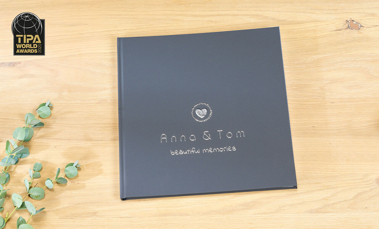 A CEWE photo book with a black cover design, personalized with the names Anna and Tom and the text 'beautiful memories'. It lies on a light wooden table with a plant in the background.