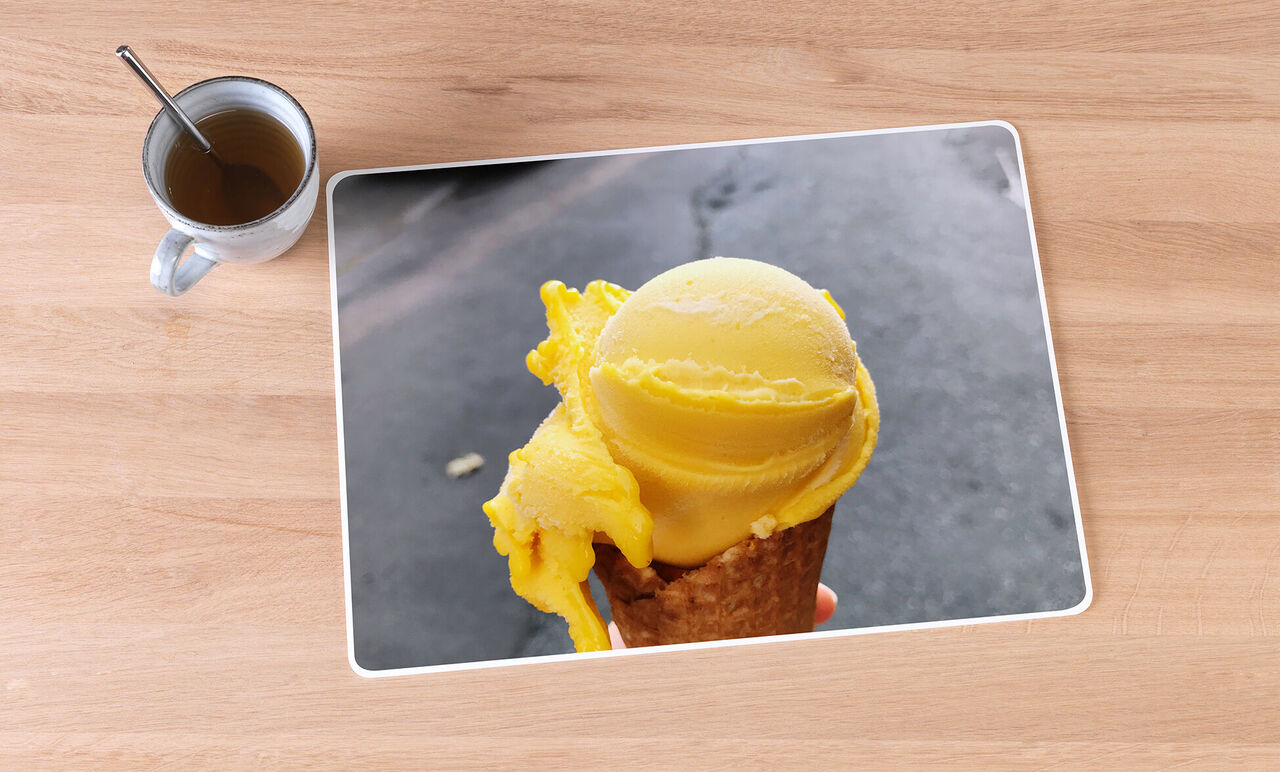 A delicious yellow ice cream on a cone, held in one hand, with a coffee next to it.