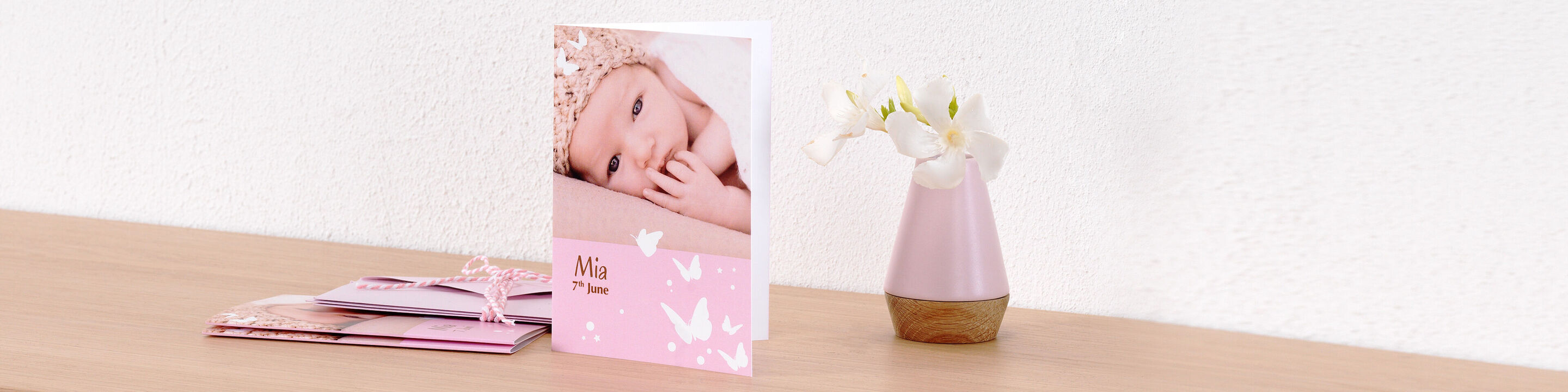 A CEWE greeting card with a smiling baby photo and delicate butterflies on a pink background, next to a blooming pot.