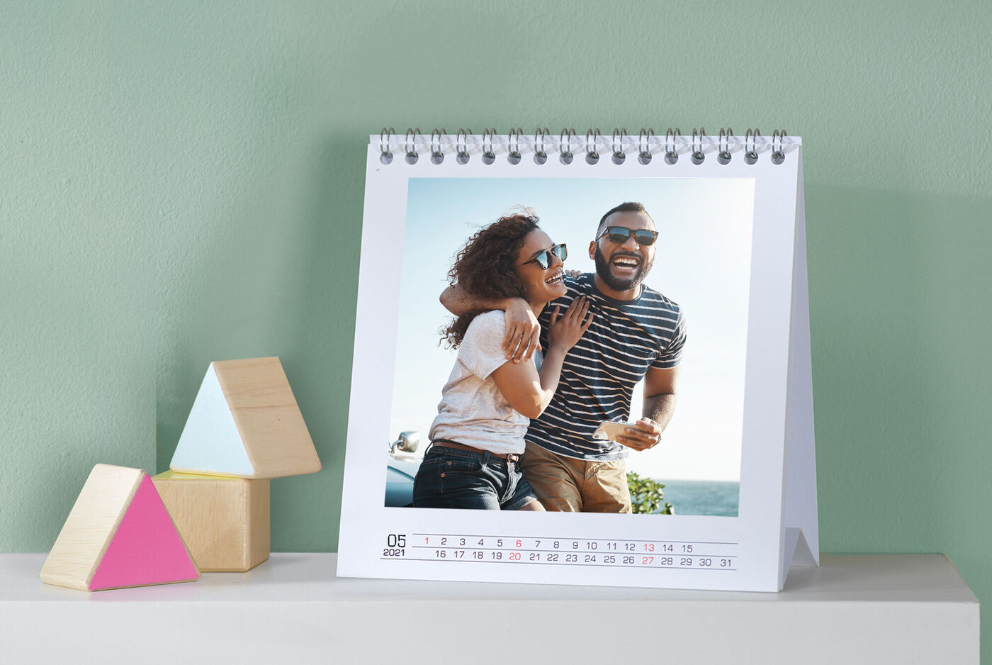 Couple smiles beside a calendar table; various geometric wooden toys are in the foreground.