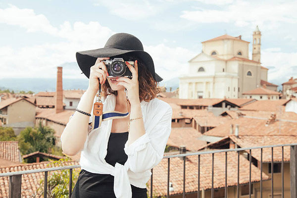A woman with a black hat and camera overlooks a city with red roofs.