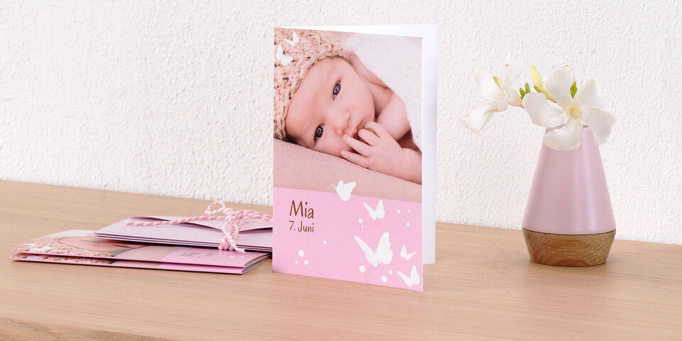 A delicate pink greeting card with a photo of a sleeping baby. The card says 'Mia 7th June' in a loving font. Beside it is a decorative vase with flowers.
