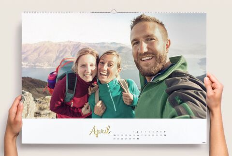 A group of three people on a mountain overlooking a landscape, holding a photo calendar for April.