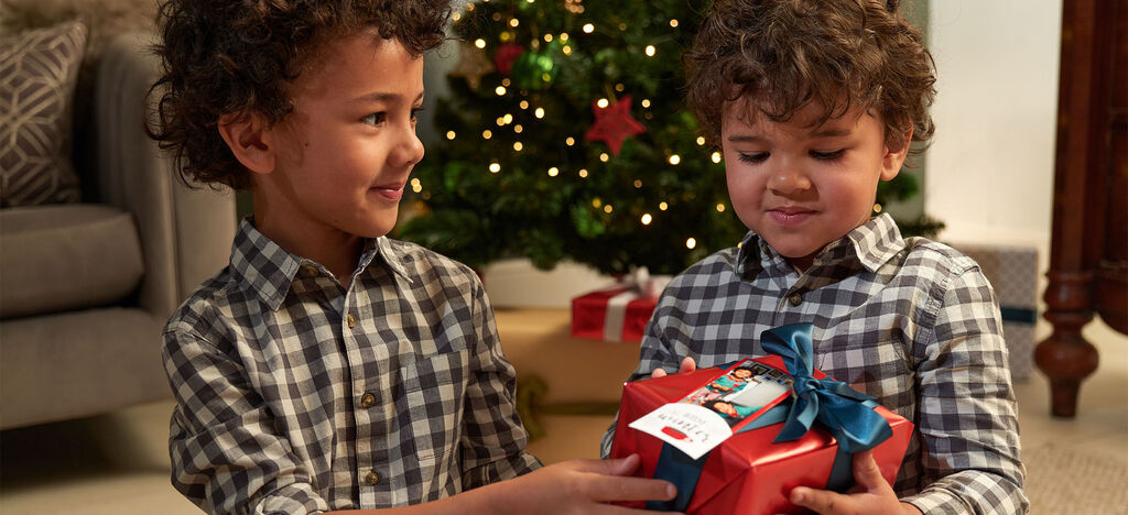 Two children exchange a red gift in front of a festively decorated Christmas tree.