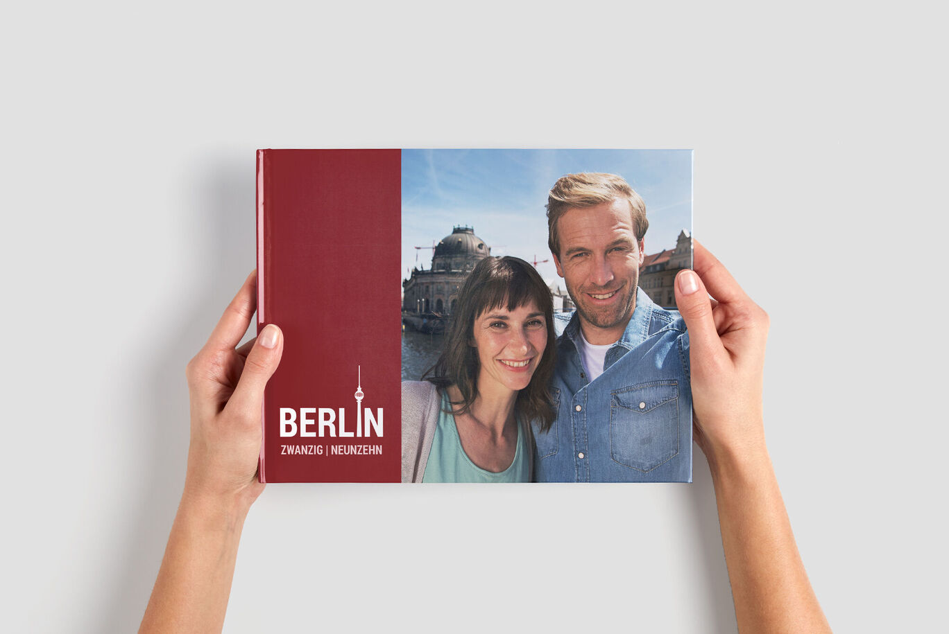 Two people holding a CEWE PHOTOBOOK titled 'Berlin' in their hands, against a backdrop of gray room