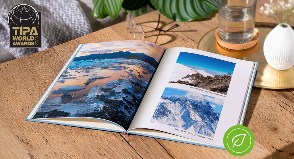 An open photo book on a wooden table showing stunning landscape photos. A glass of water and a plant are also visible.