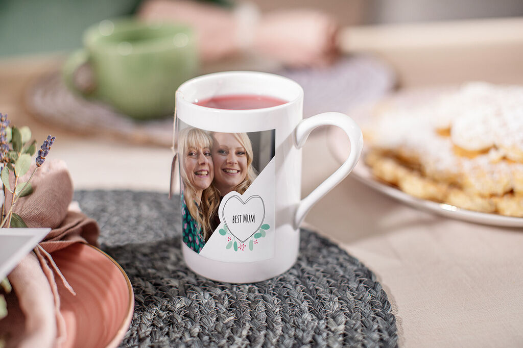 Person holds a personalized mug with the print 'BEST MOM'. Nearby is a plate of treats and a green mug.
