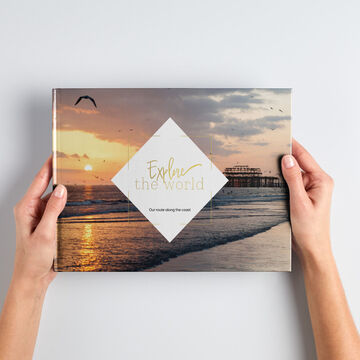 A person holds a CEWE photo book with a sunset at the beach on the cover.