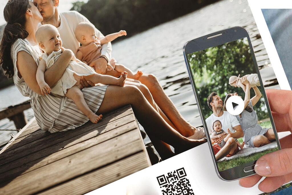 A family enjoys a day by the water. Parents sit on a wooden platform holding their babies. A phone shows a video of happy moments.