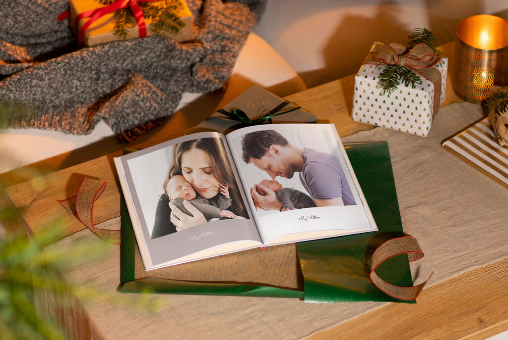 An open photo book on a table with gifts and decorations. It shows a personal photo page with a grandparent-grandchild picture.