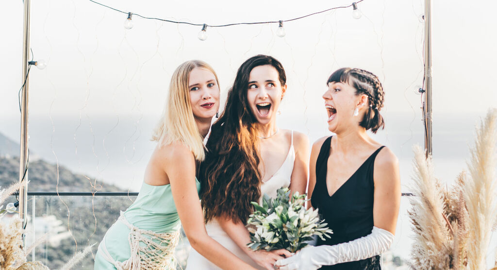 Three women in festive attire embrace lovingly while holding a bridal bouquet.