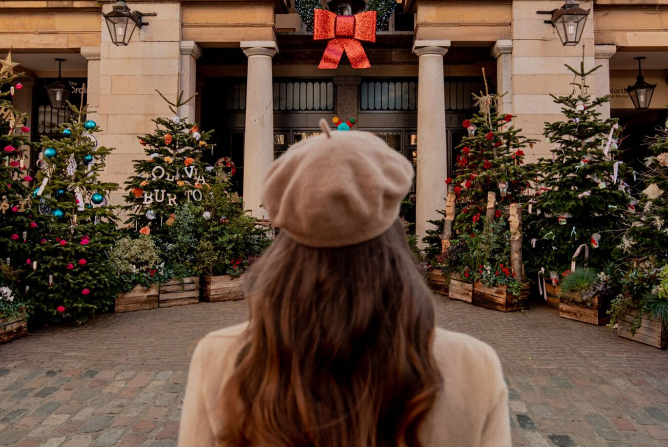 Woman with long wavy hair stands in front of a festively decorated Christmas scene with trees and big colorful ornaments.