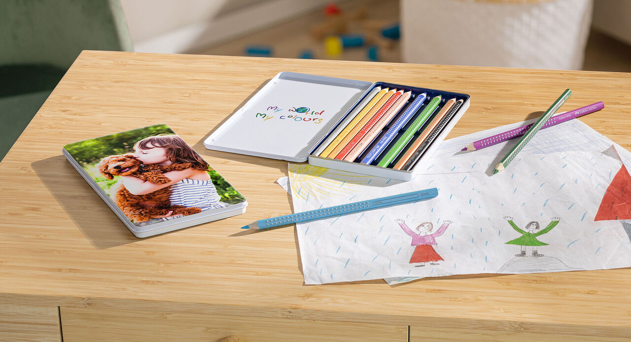 A CEWE photo book lies on a wooden table next to colored pencils and children's drawings