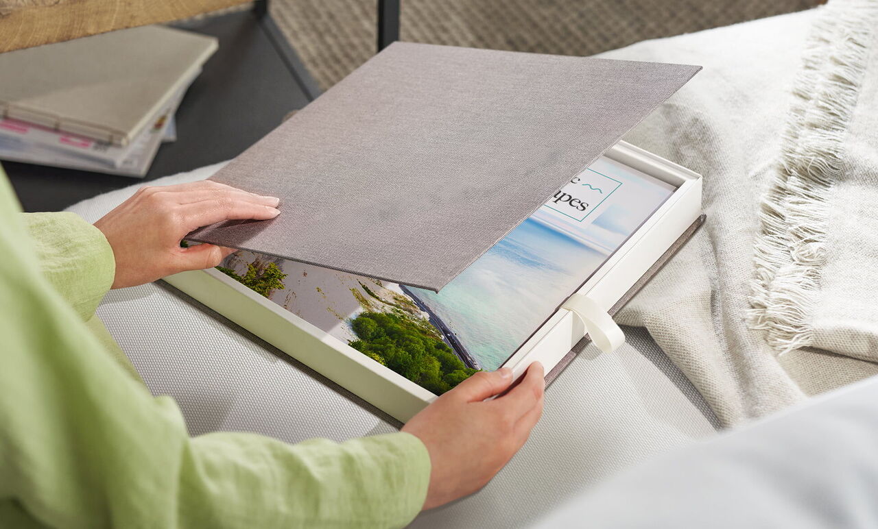 A person opens a CEWE photo book with pictures from vacation. The images show beautiful landscapes and memories.