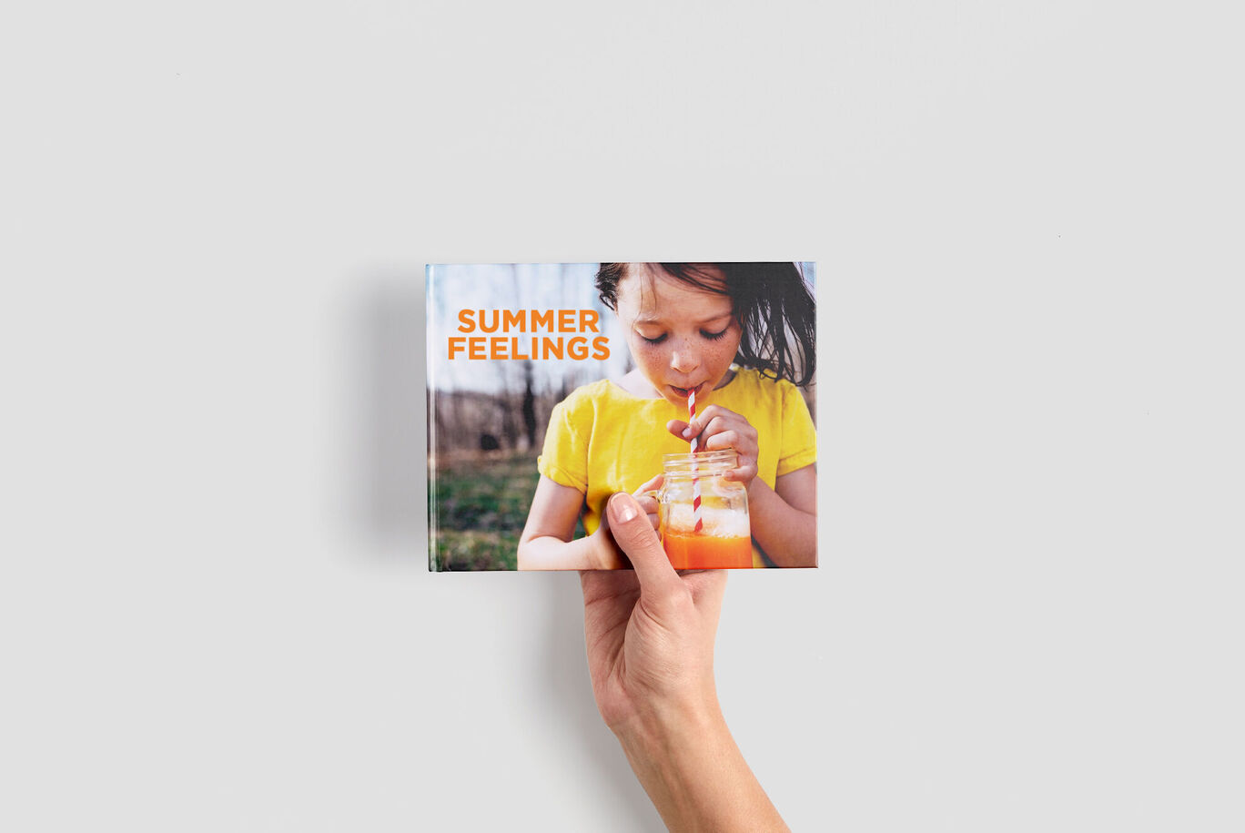 A hand holds the CEWE PHOTOBOOK "Summer Feelings" showing a person with a drink.