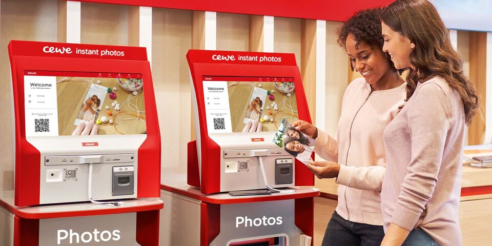 Two women use a CEWE machine for instant pictures. One woman shows a screen with photos.