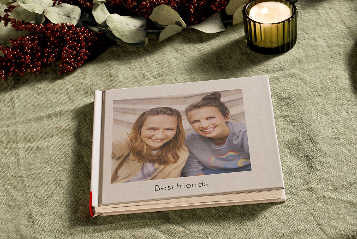 A CEWE PHOTO BOOK on a table, surrounded by decorative elements. It shows a community picture of two friends with 'Best friends' on the cover.