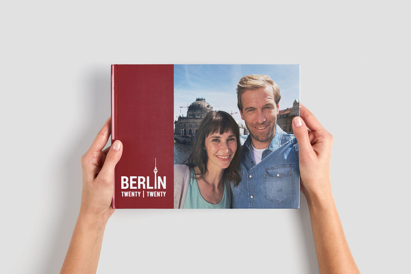 A couple holds a CEWE PHOTOBOOK in their hands, showcasing a travel experience in Berlin. The surroundings are sunny and lively.