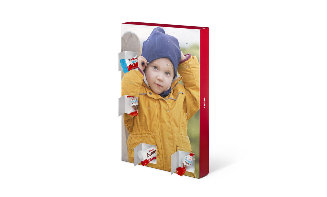 A child in a yellow raincoat stands joyfully in front of a large red wall with compartments for surprises. It reaches for a treat.