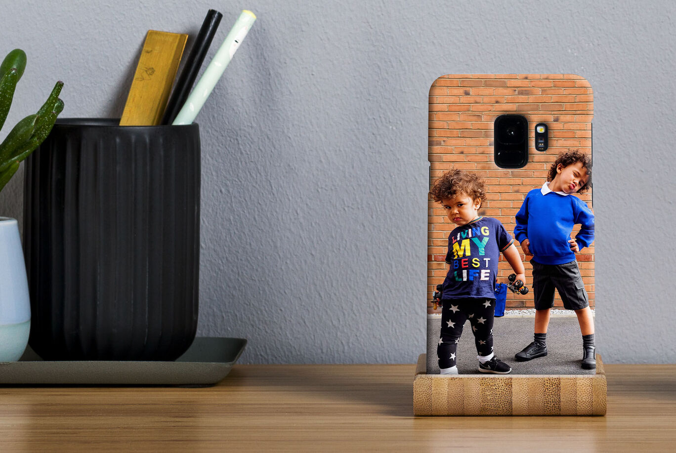 Kid in a colorful t-shirt stands next to another kid in blue sweater, posing. Background shows a modern living space with plants.