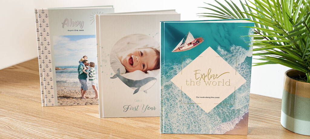 Three individually designed CEWE photo books on a table, featuring a family at the beach, a baby with a dolphin, and a boat on the water.
