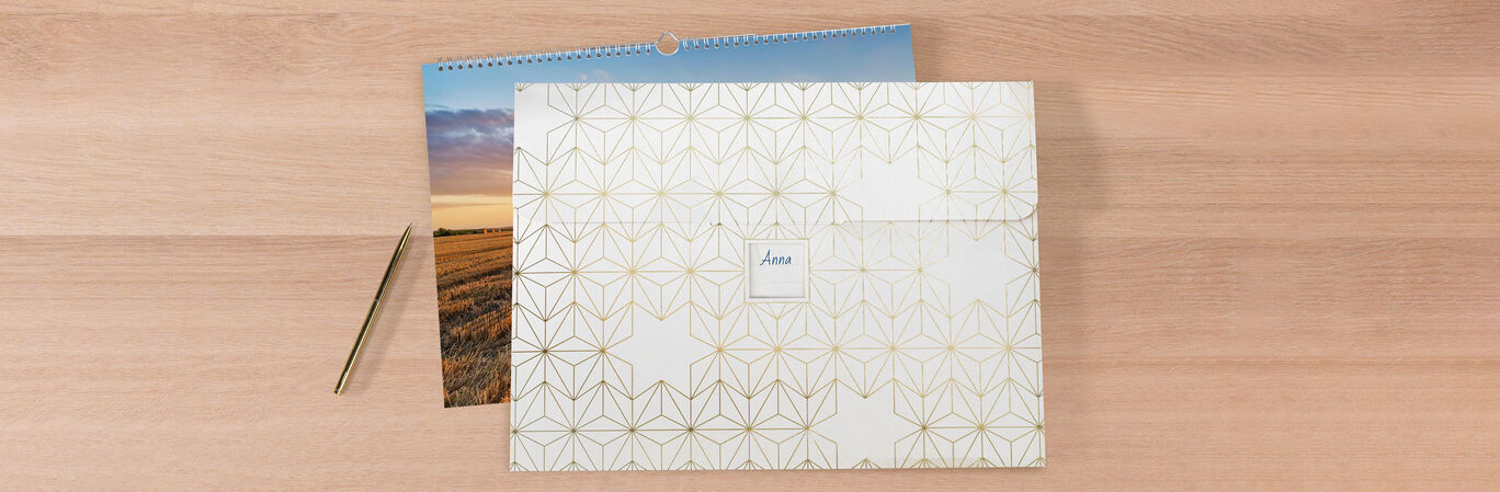 A beautifully designed CEWE photo calendar lies on a table, alongside an elegant folder with a simple design. In the background, part of the calendar shows a stunning sunset.