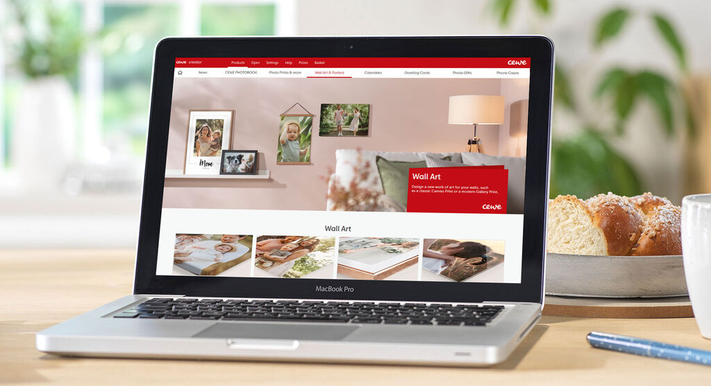 Laptop showing the CEWE wall picture page with photos and templates, surrounded by a cozy living atmosphere