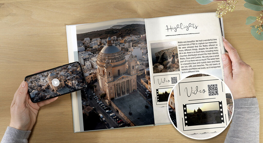 A person holds a smartphone showing a video while looking at a photo book with city views, featuring QR codes