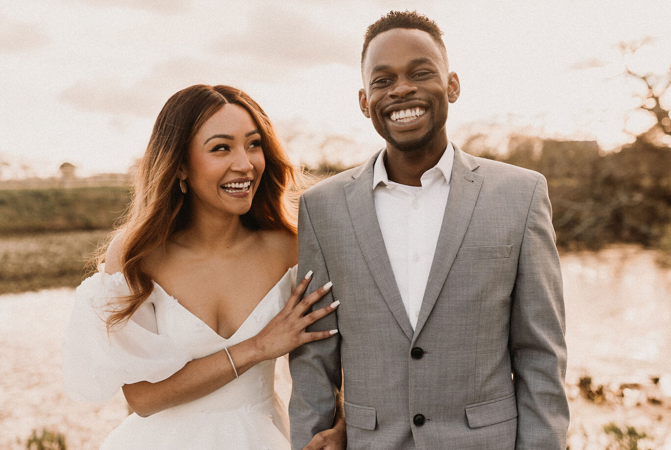 A couple stands at the shore of a body of water smiling at each other. They wear elegant clothing, the man in a gray suit and the woman in a white wedding dress. The surrounding nature glows in warm colors, creating a romantic atmosphere.