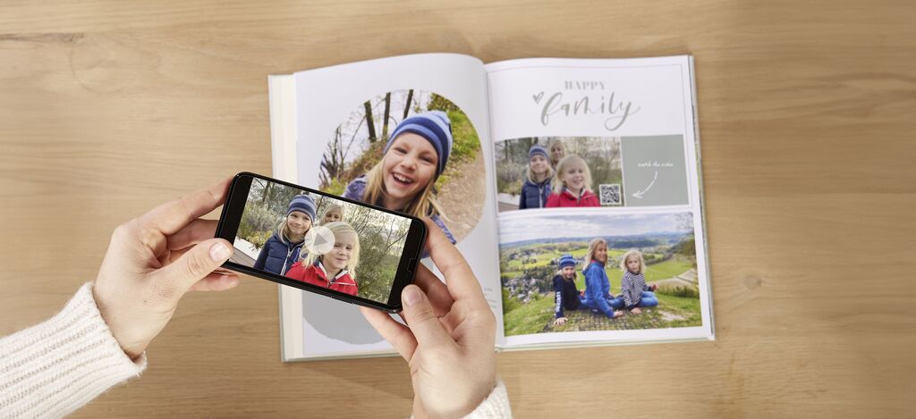 A person holds a phone showing photos while viewing an open photo book with family images