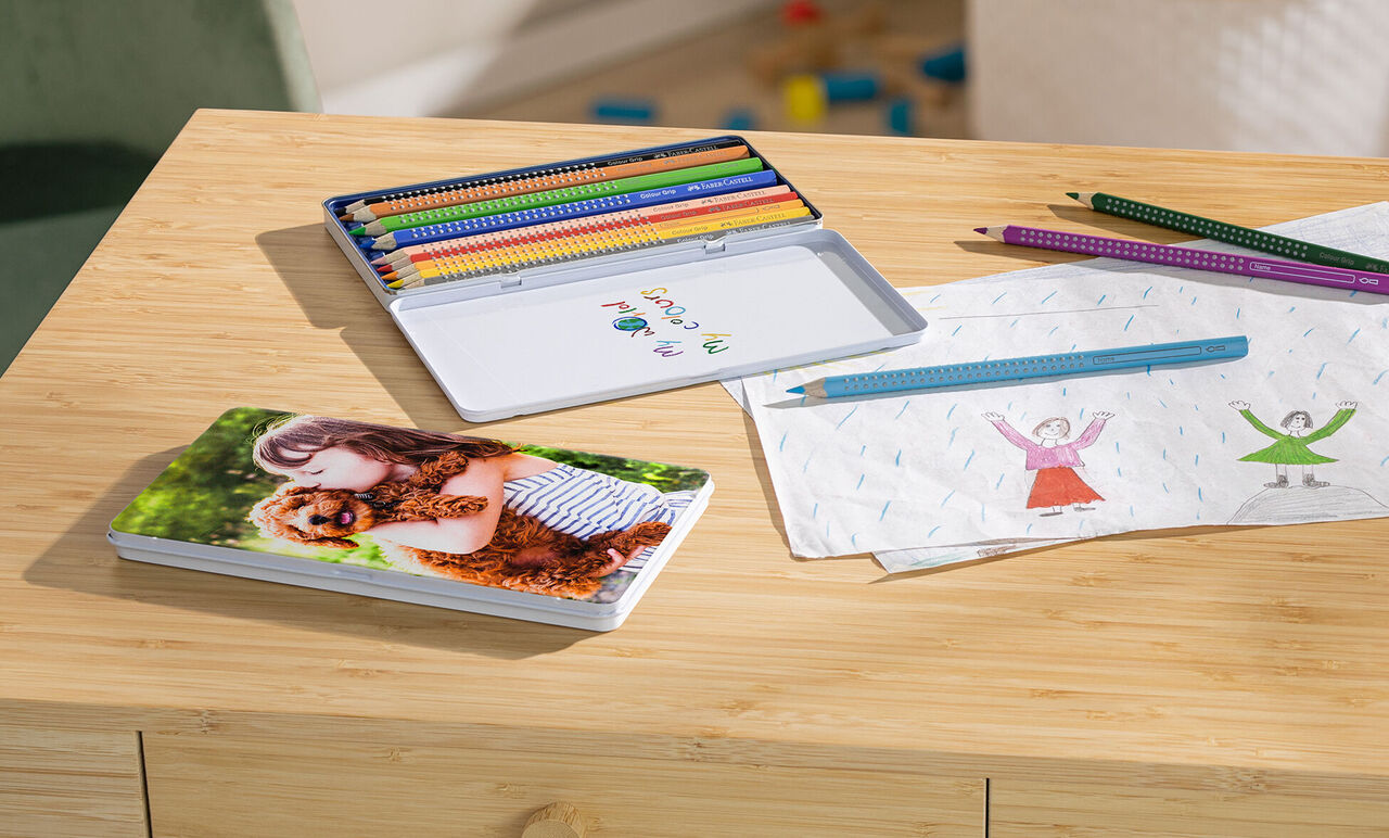 A CEWE photo book with a smiling couple lies on a table next to colorful pencils and drawings.
