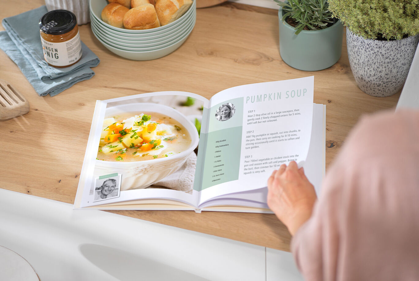 A person is flipping through a recipe book on a kitchen counter. It shows a creamy pumpkin soup with fresh herbs and vegetables.