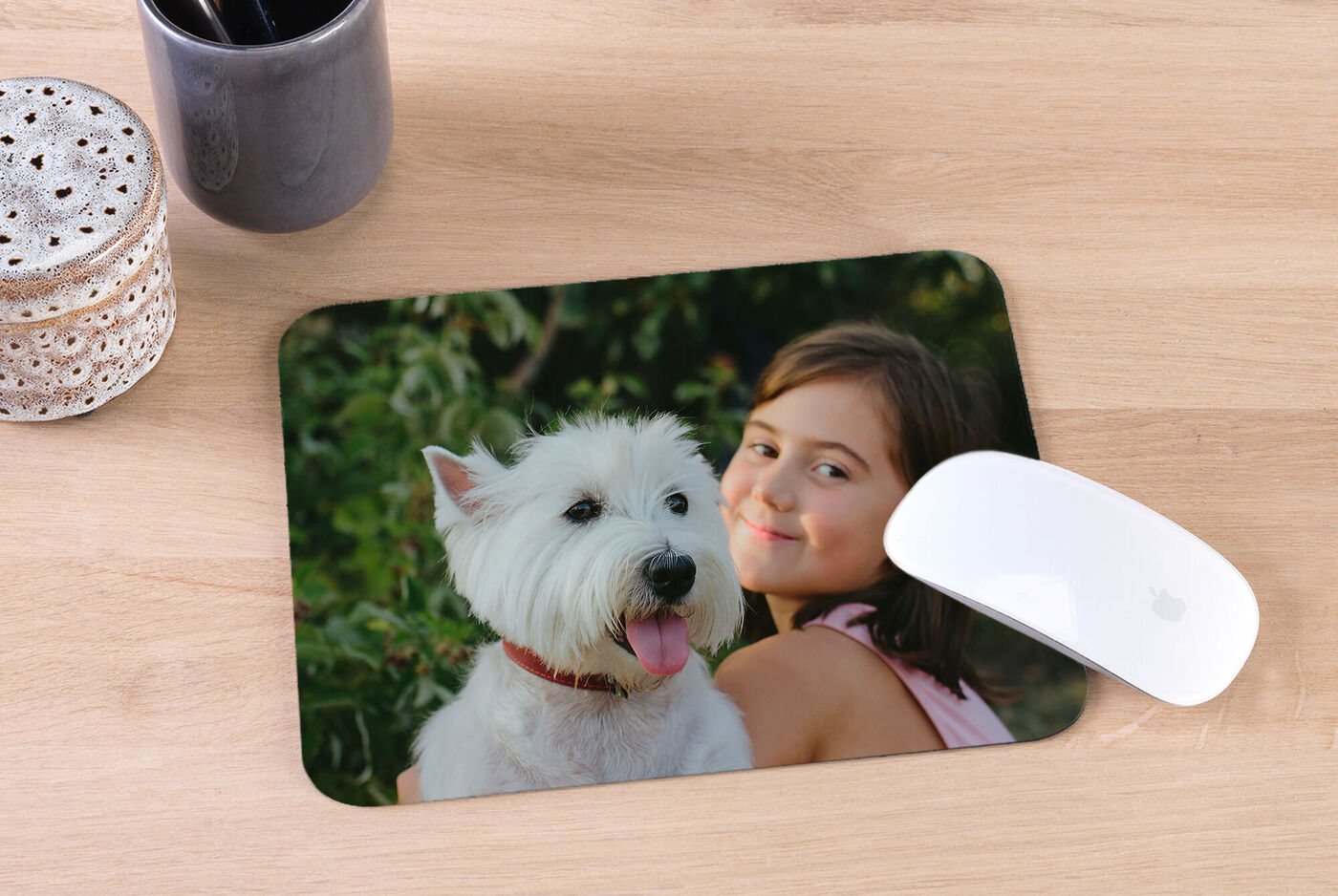 A boy or girl hugs a white dog outdoors and smiles, surrounded by green plants.