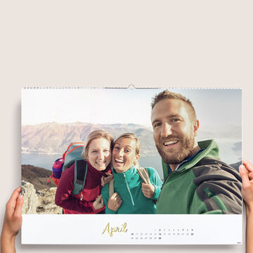 Two people holding a CEWE photo calendar pointing at April, mountains in the background.