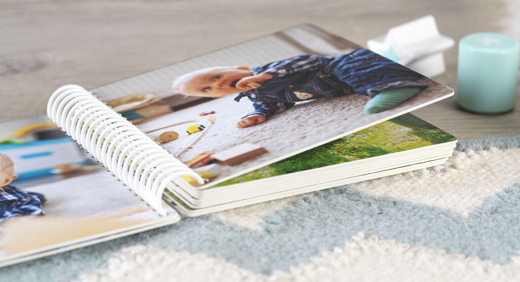 A CEWE photo book lies on a table, with a baby looking at a colorful toy page.
