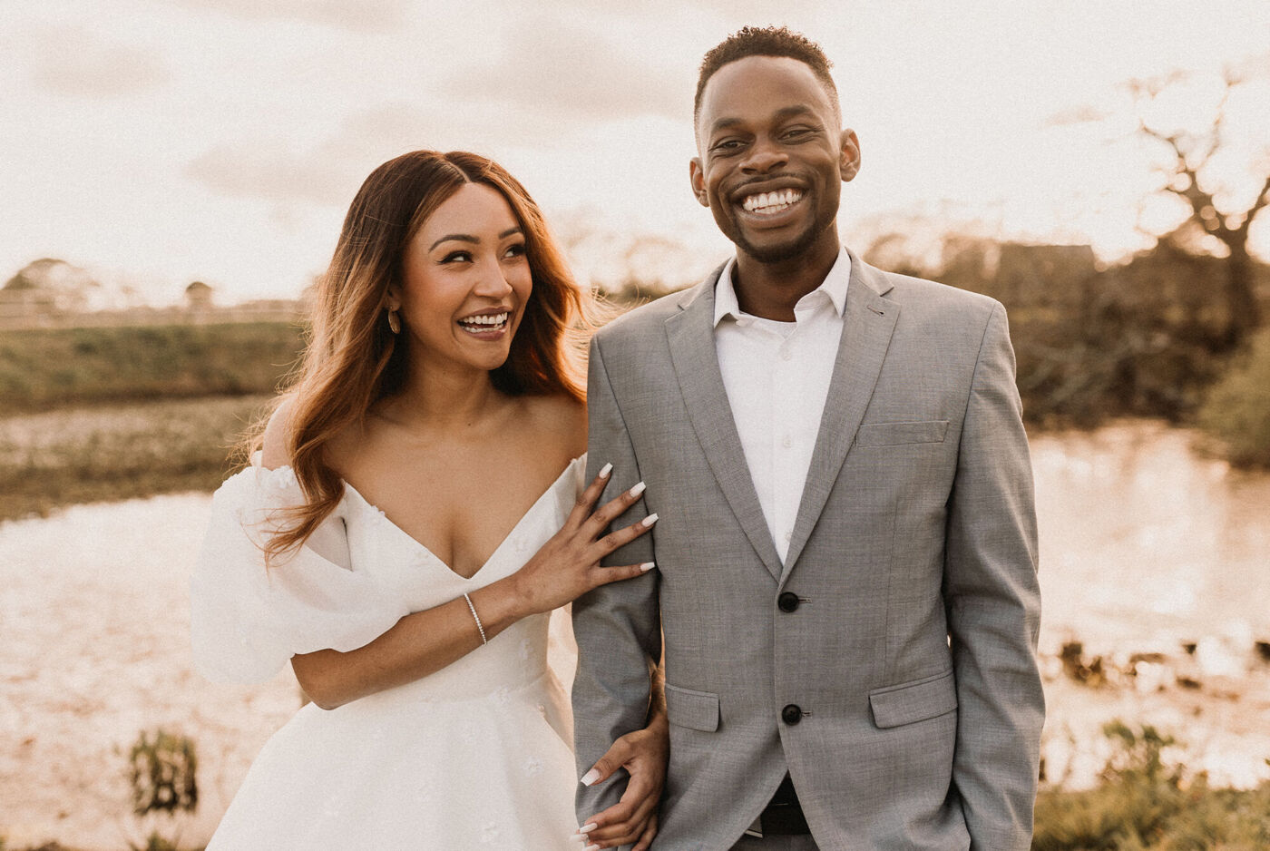 A couple in elegant attire stands by a body of water, surrounded by nature.