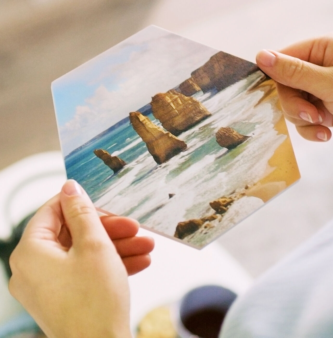 A person holds a hexagonal photo card of the famous Twelve Apostles on the Australian coast. The ocean and rocks are visible.