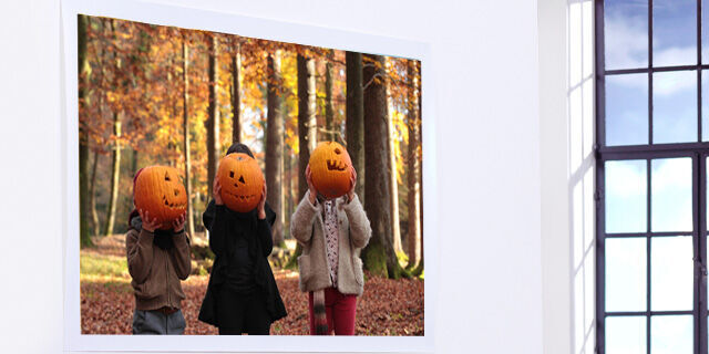 Three children in a forest wearing pumpkin head masks in front of colorful autumn leaves.