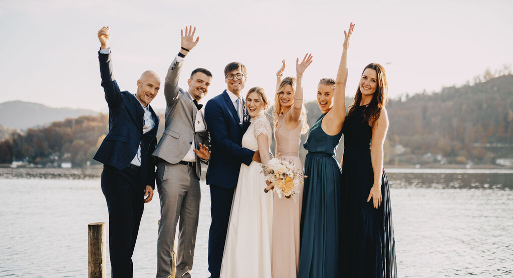 Friends joyfully celebrate by the lake, dressed elegantly, smiling and waving.