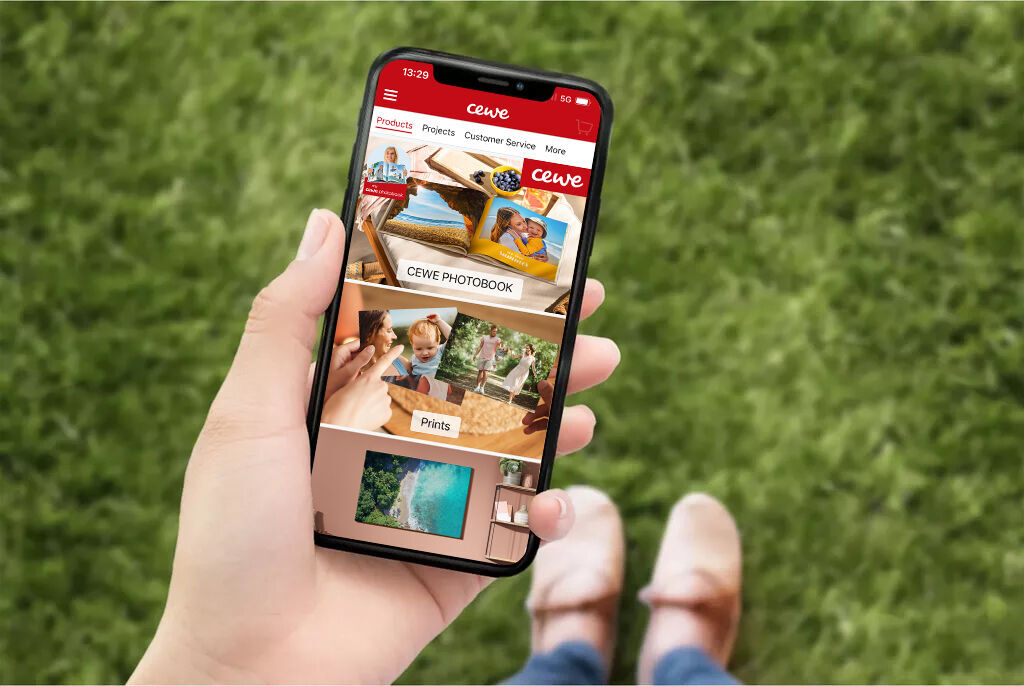 A hand holds a smartphone with the CEWE photo book app open on green grass