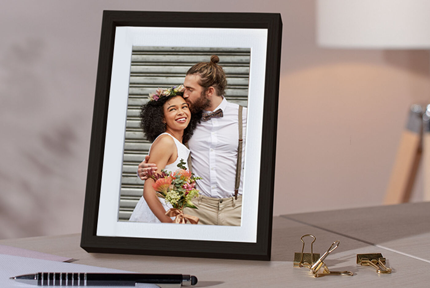A loving couple stands close together sharing an intimate moment. They wear festive clothing, embracing against a soft gray wall backdrop, with flowers adorning their hair.