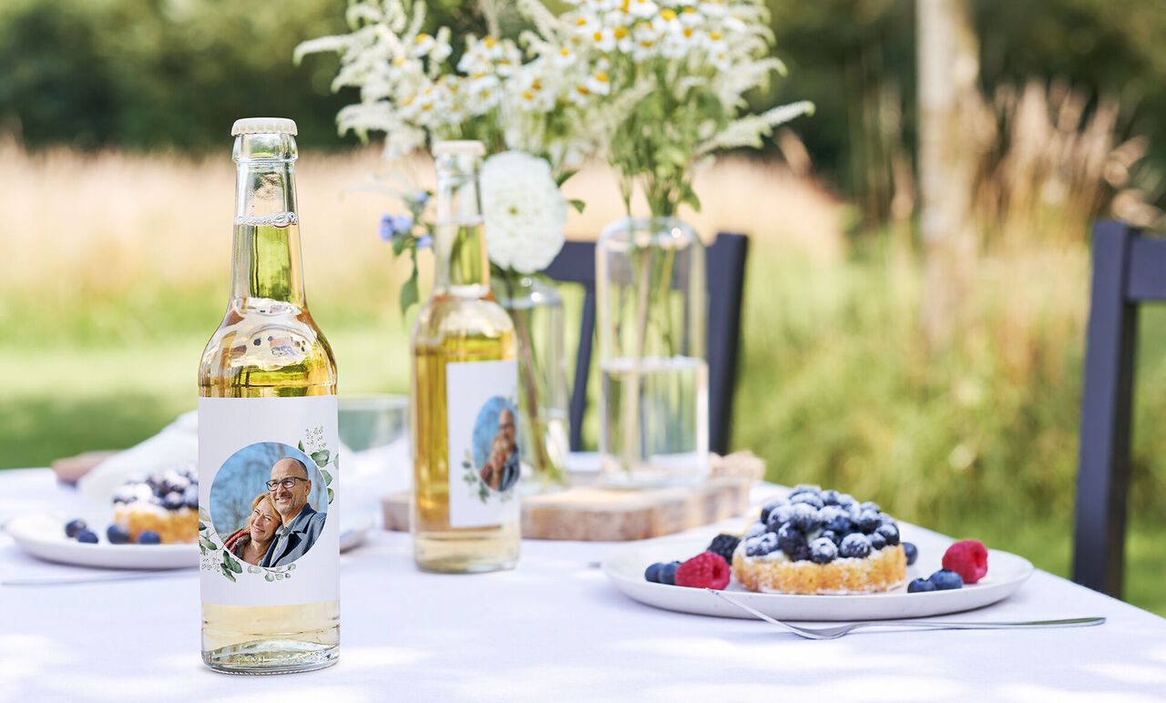 Two bottles with personalized labels on a table set with berries and a cake.