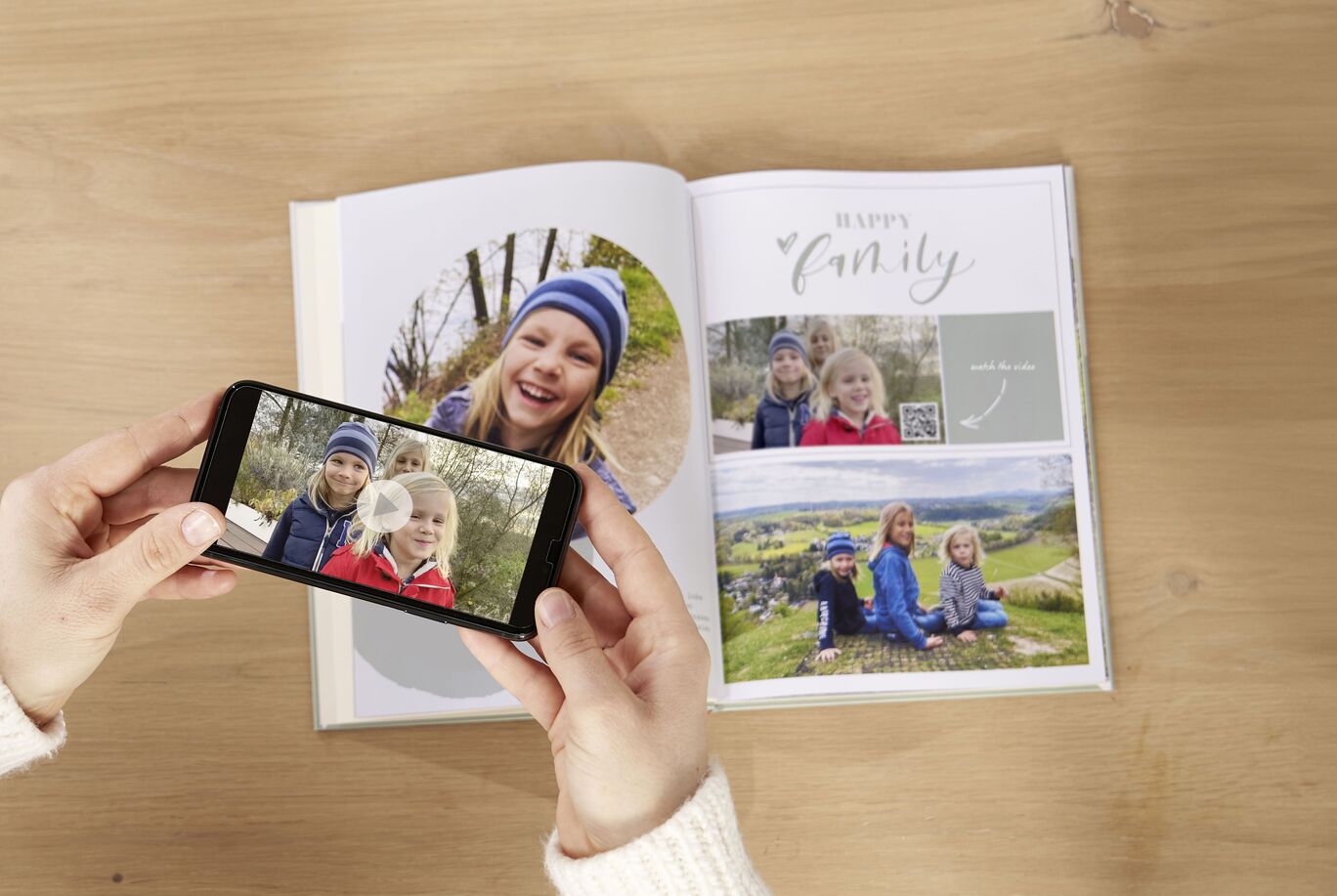 A person holding a smartphone and photographing a page from a CEWE PHOTOBOOK. The page shows two children happily sitting outside.
