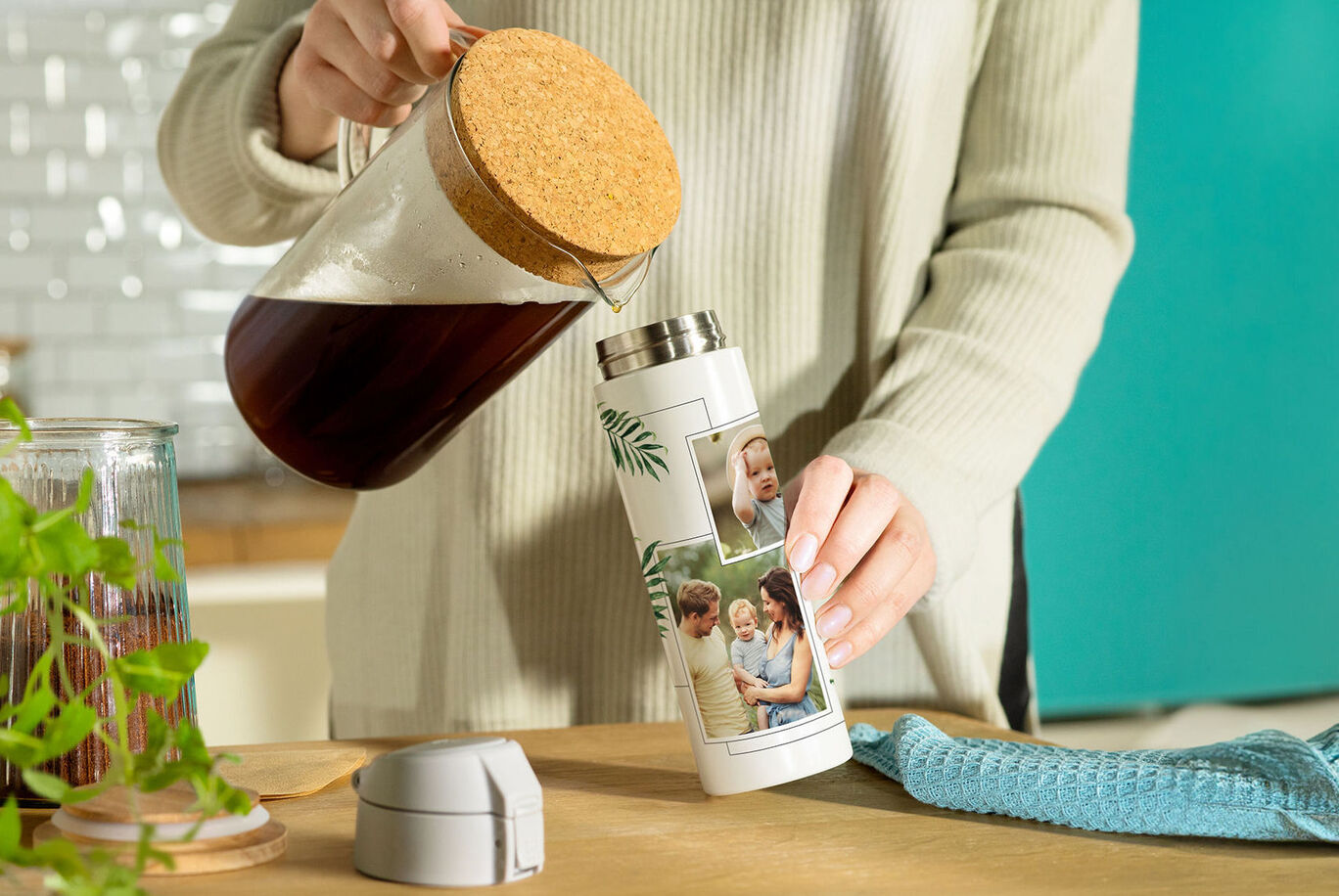 A person pours coffee from a pot into a personalized thermal mug with photos. A towel and a plant are on the table.
