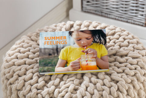 A child holds a glass with orange drink, wearing a yellow shirt. A CEWE photo book titled 'SUMMER FEELINGS' is on the table.