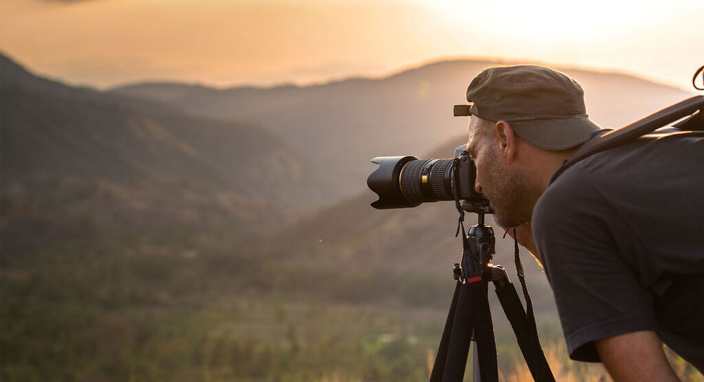 A photographer stands with a camera on a tripod in a breathtaking mountain landscape. The sun shines, creating a warm atmosphere.