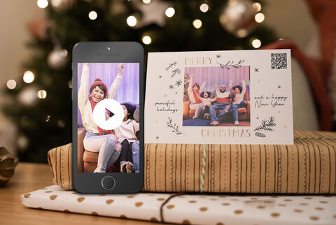 A person holds a phone showing a video, while another person sits on the couch showing joy surrounded by gift wrap.